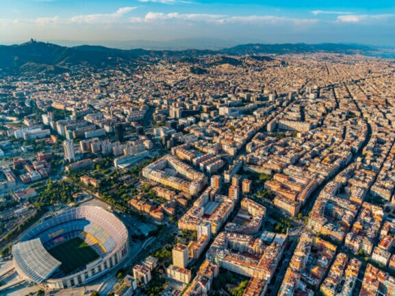 Stadtpanorama von Barcelona mit Fußballstadion aus der Luftperspektive, im Hintergrund eine Hügelkette und Meer.