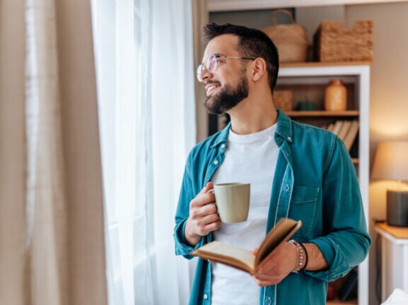 Ein Mann im Jeanshemd mit Buch und Kaffeebecher in den Händen schaut entspannt aus einem Fenster mit Vorhängen.