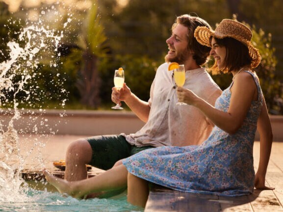 Eine Frau und ein Mann sitzen am Rand eines Pools mit einem Getränk in der Hand und spritzen mit ihren Füßen.