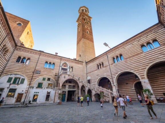 Personen in einem italienischen Palastinnenhof mit gestreifter Fassade und Außentreppe unter einem Turm vor blauem Himmel.