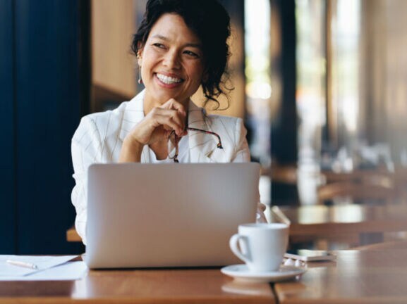 Eine Frau sitzt zufrieden lächelnd an einem Laptop an einem Tisch mit Kaffeetasse in einem Café.