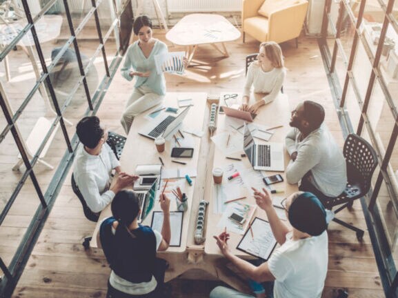 Vogelperspektive in einem Büro mit großem Tisch, an dem mehrere Personen mit Laptop sitzen und miteinander sprechen.