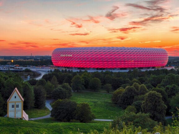 Panoramaaufnahme eines pink leuchtenden Fußballstadions unter orange gefärbtem Himmel bei Sonnenuntergang, im Vordergrund Wiesenfläche mit Wald und Kapelle.
