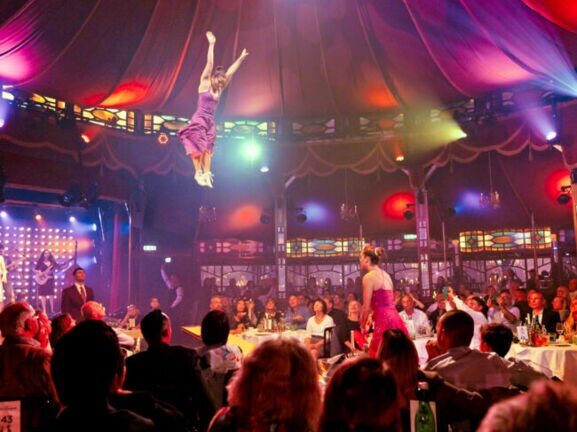 Personen sitzen beim Dinner an Tischen in einem Spiegelzelt während einer Show mit Akrobatinnen.