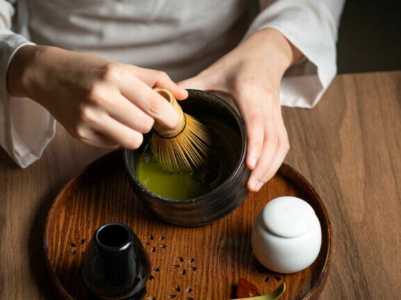 Nahaufnahme von zwei Händen bei der Zubereitung eines grünen Tees mit einem Bambusbesen in einer dunklen Keramikschale auf einem Holztisch.