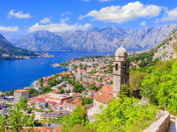 Blick auf die Stadt Kotor mit Häusern und Kirchturm, im Hintergrund Berge und eine Bucht.
