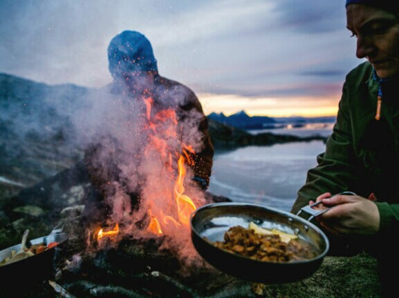 Zwei Personen bereiten Speisen in Pfannen über einem Feuer an einer schroffen Felsküste zu bei Abenddämmerung.