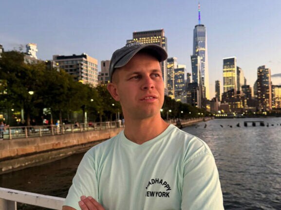 Porträt von Benjamin Diedering mit Baseballcap vor der Skyline New Yorks am Wasser.