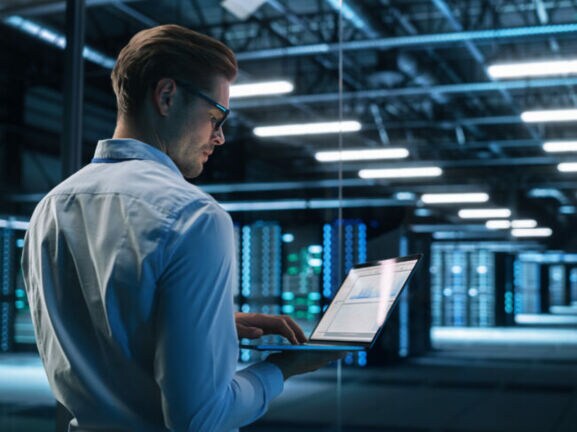 Profil eines Mannes im Hemd, der mit einem aufgeklappten Laptop in einem Rechenzentrum steht.