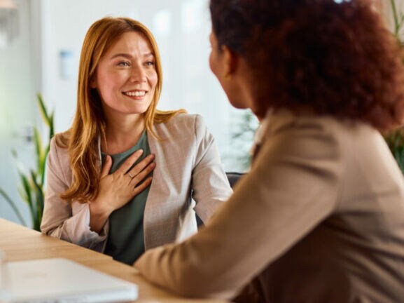 ine Frau im Blazer hat eine Hand auf die Brust gelegt und lächelt eine weitere Frau an, beide sitzen an einem Tisch, auf dem ein Tablet und ein Wasserglas stehen.