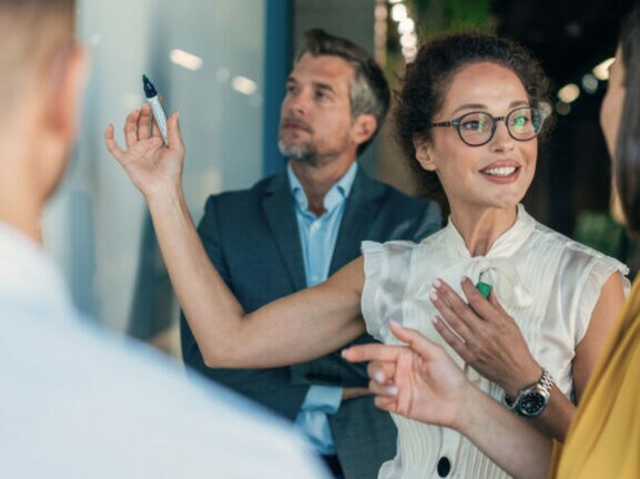 Vier Personen in Business-Kleidung stehen vor einer Wand und kommunizieren, eine Frau hält einen Stift in der Hand hoch.