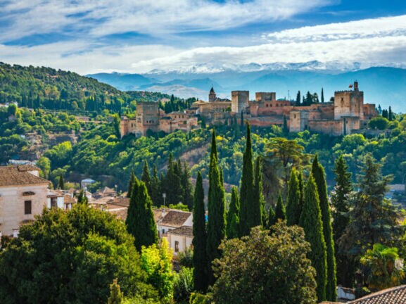 Blick auf einzelne Häuser und die von Wald umgebene Alhambra, im Hintergrund Berge.