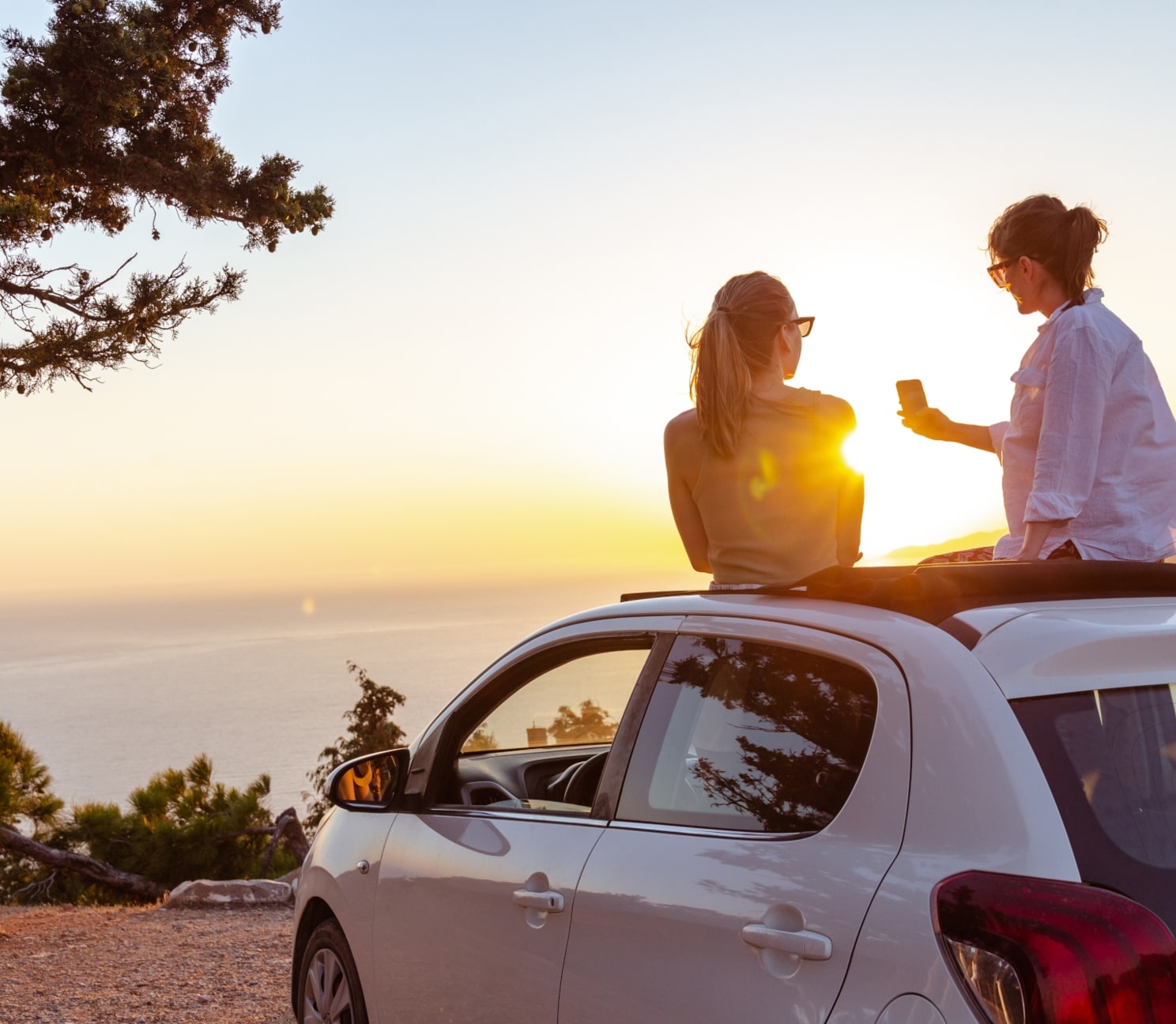 Rückansicht zweier junger Frauen auf dem Dach eines weißen Autos an einer Meeresküste bei Sonnenuntergang.