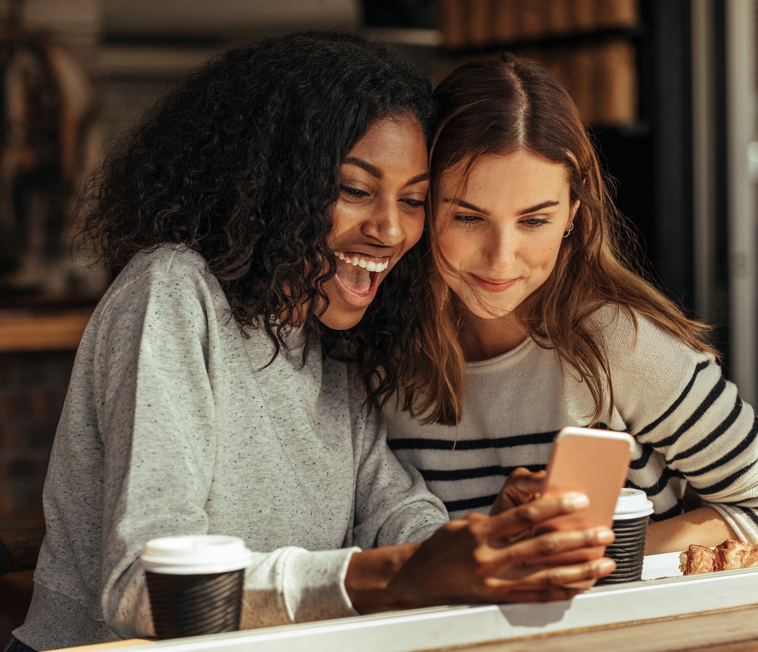 Zwei Frauen schauen lachend im Café gemeinsam auf ein Smartphone.