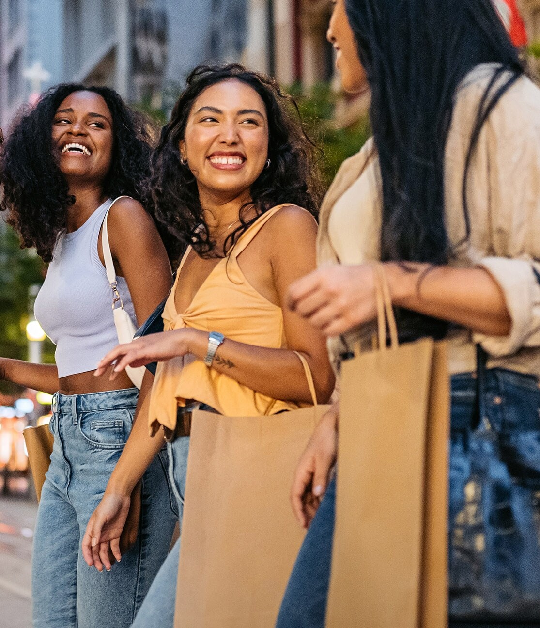 Drei sommerlich gekleidete Frauen mit Einkaufstüten in den Händen laufen gut gelaunt nebeneinanderher auf einer Straße im Stadtzentrum.