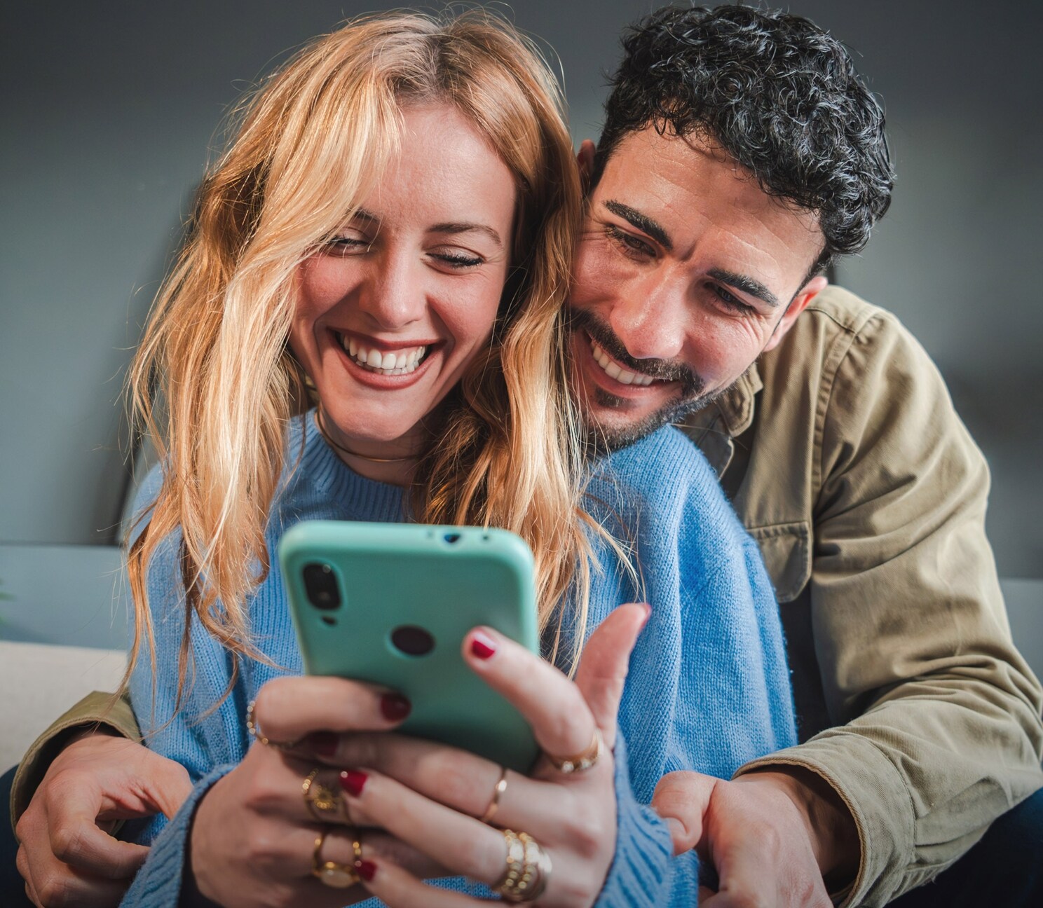 Eine Frau und ein Mann sitzen eng umschlungen auf einem Sofa und blicken lachend auf ein Smartphone in ihren Händen.