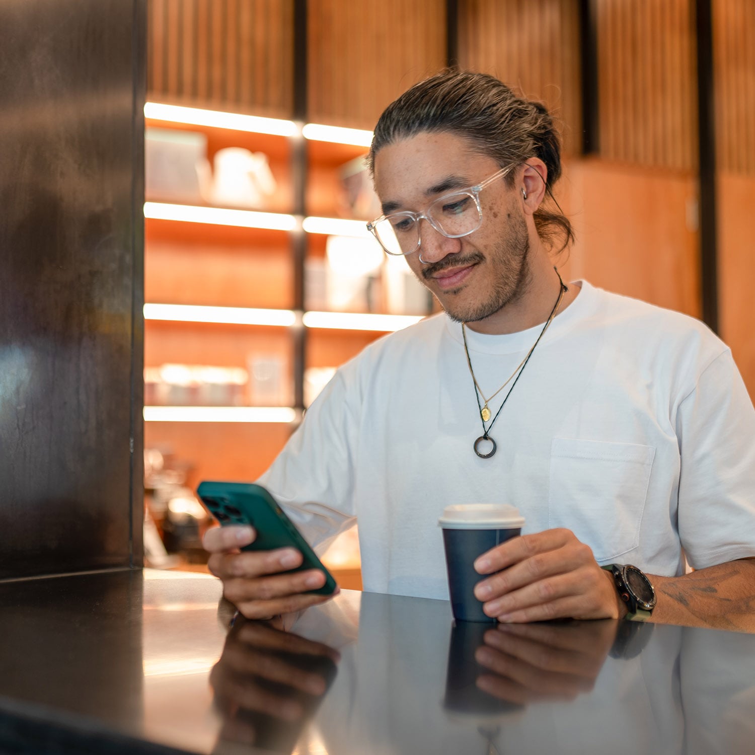 Ein stylischer Mann mit Zopf im weißen T-Shirt steht mit einem Kaffeebecher an einem Tresen in einem Geschäft und schaut auf sein Smartphone.