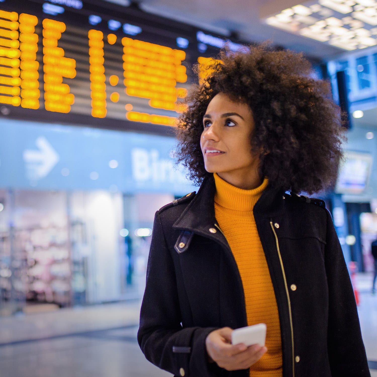 Eine lächelnde Frau in Lederjacke mit Smartphone in einem Flughafenterminal.