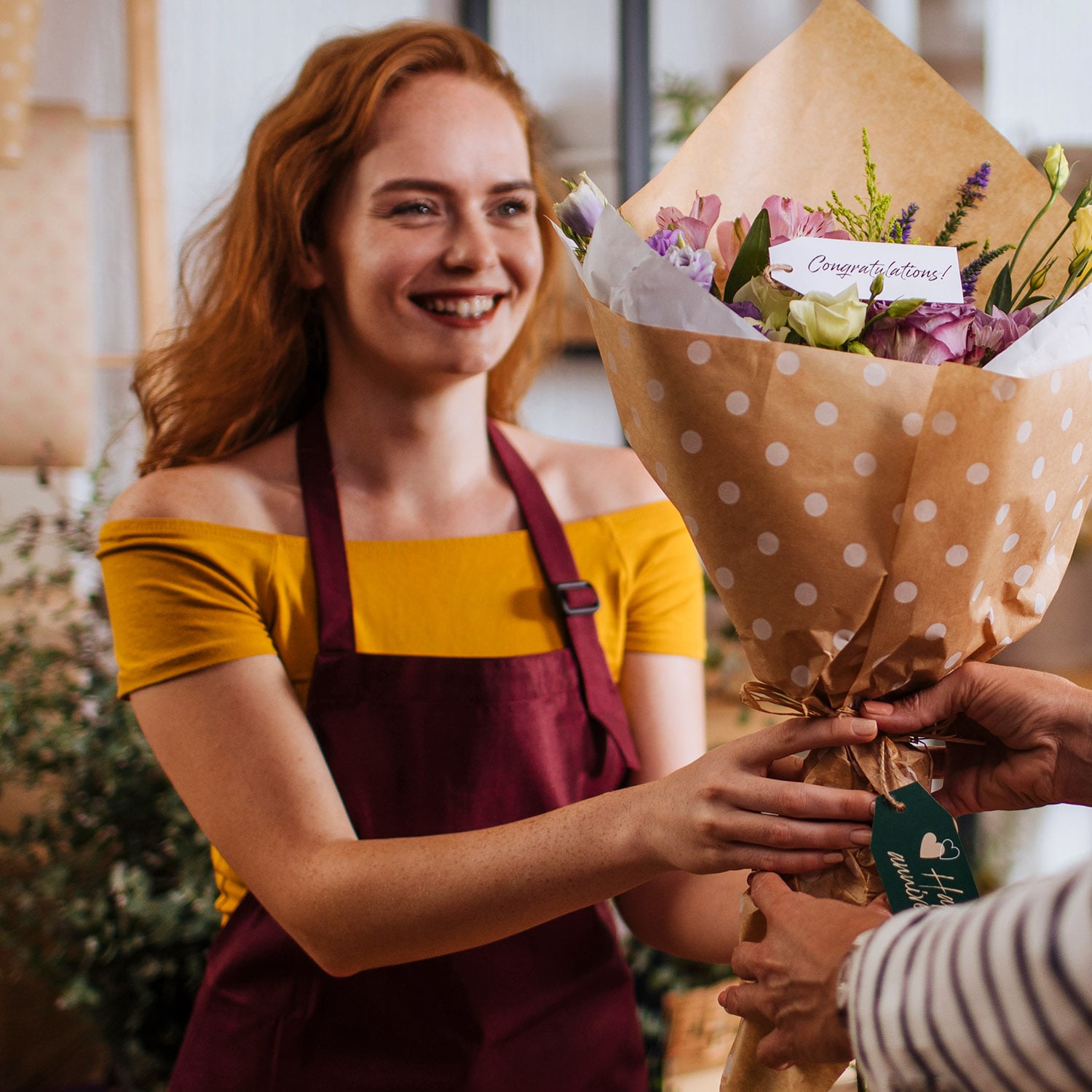 Eine lächelnde, rothaarige Verkäuferin mit Schürze überreicht einen Blumenstrauß.