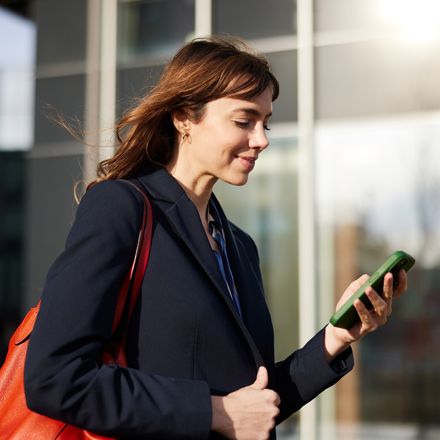 Eine elegante Frau im Blazer schaut zufrieden auf ihr Smartphone in urbaner Umgebung.