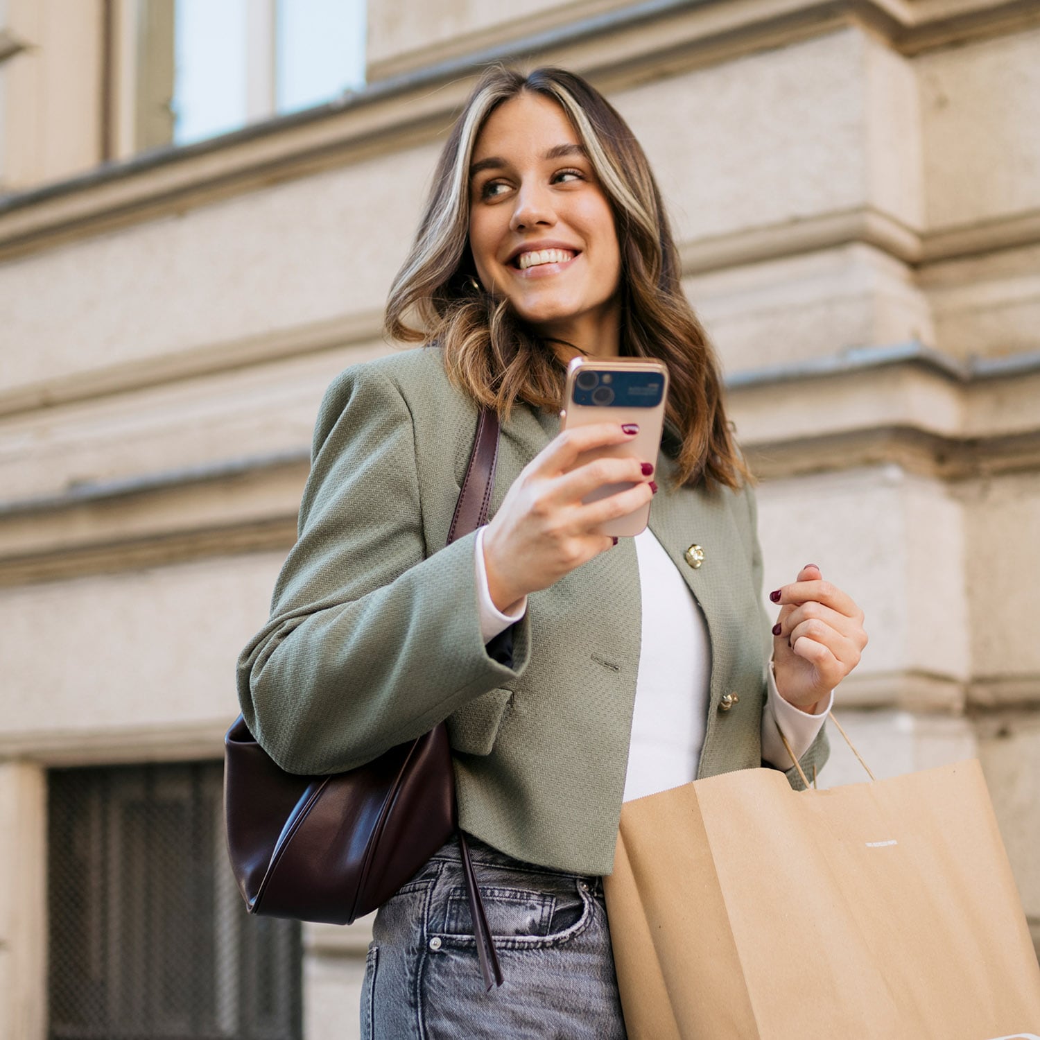 Eine junge, elegant gekleidete Frau mit Einkaufstüte und Smartphone in den Händen steht lächelnd vor einer Hausfront.