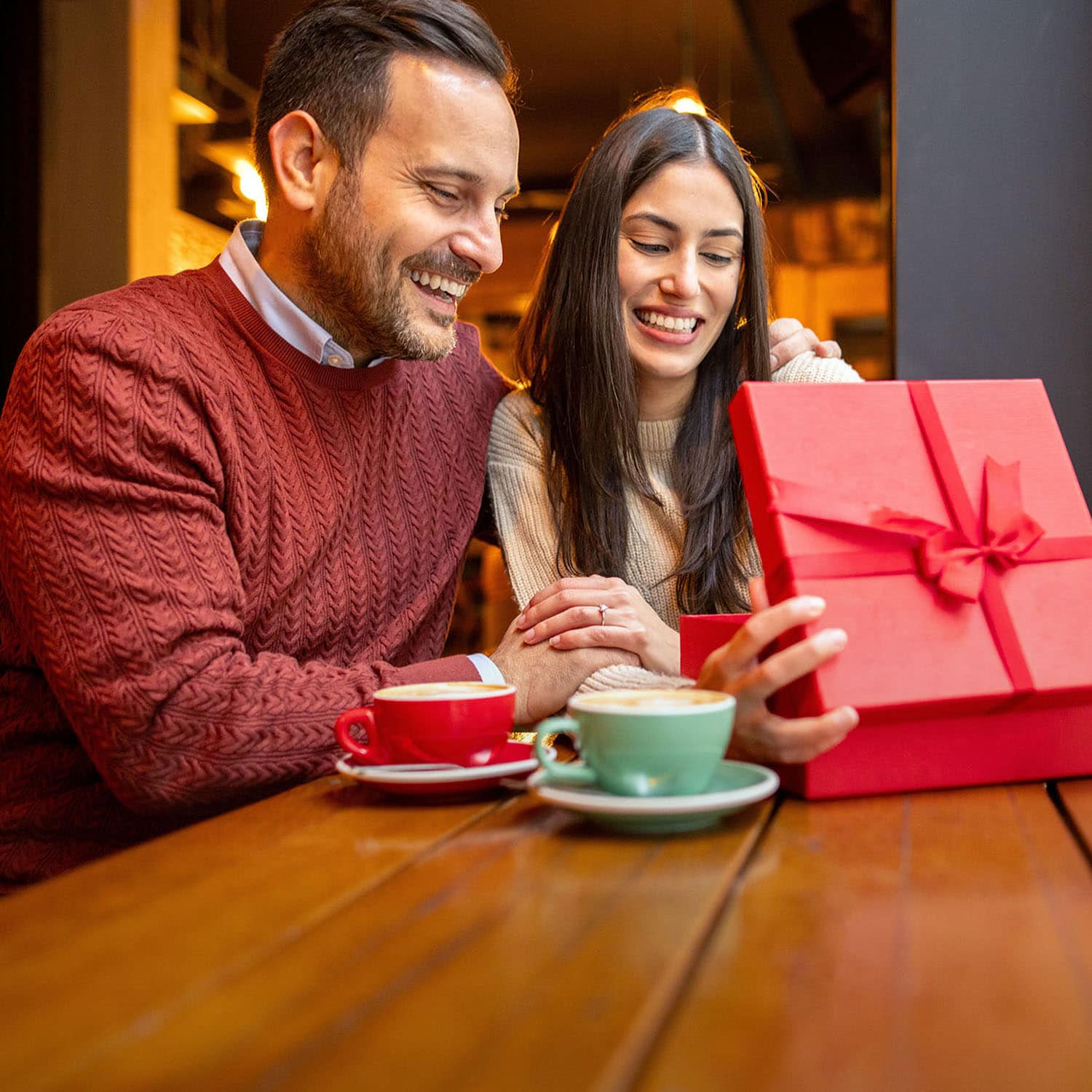 Ein lachendes Paar sitzt händchenhaltend beieinander und schaut gemeinsam in eine rote Geschenkbox mit roter Schleife an einem Tisch mit Cappuccinotassen einem Café.