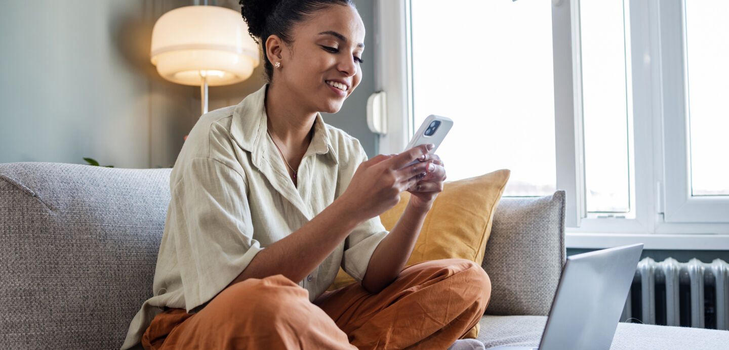 : Eine Frau sitzt im Schneidersitz auf einer Couch und schaut lächelnd auf ihr Smartphone. Vor ihr steht ein Laptop.