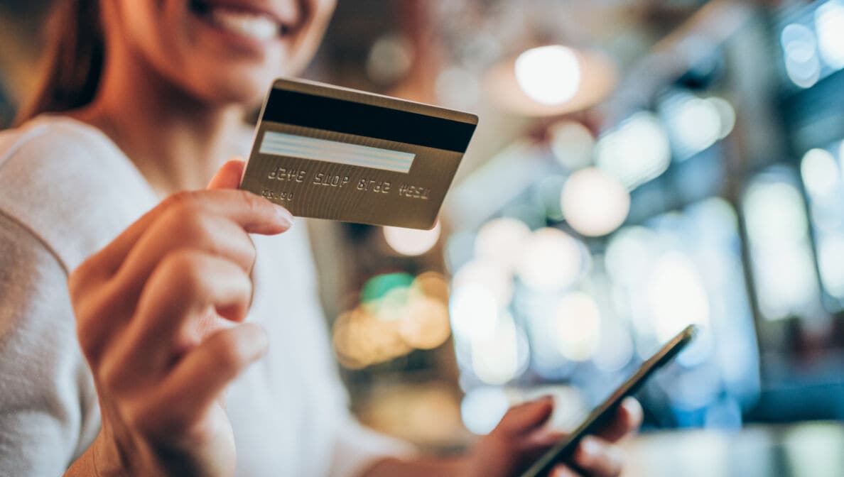 Eine lachende Frau hält ihr Smartphone und eine goldene Kreditkarte in ihren Händen