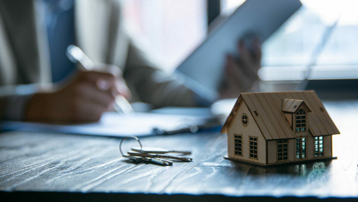 Ein Holzhausmodell auf einem Tisch mit einem Schlüsselbund daneben. Im Hintergrund sitzt eine Frau mit Dokumenten