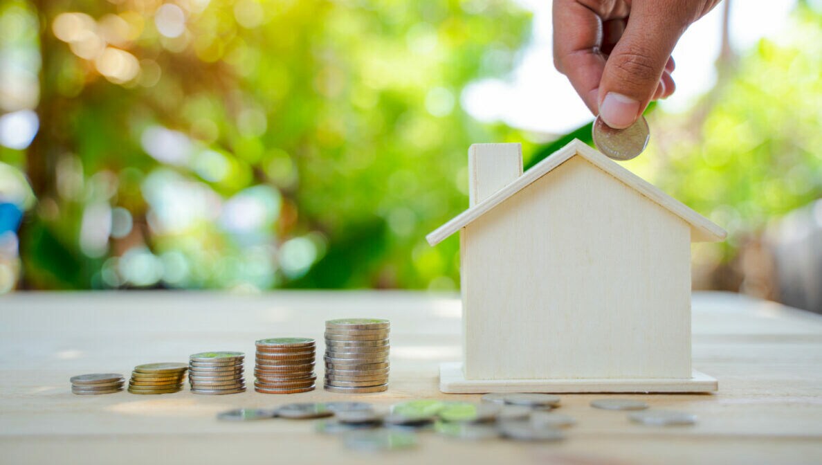Nahaufnahme einer Hand, die Münzen in eine Spardose in der Form eines Hauses steckt