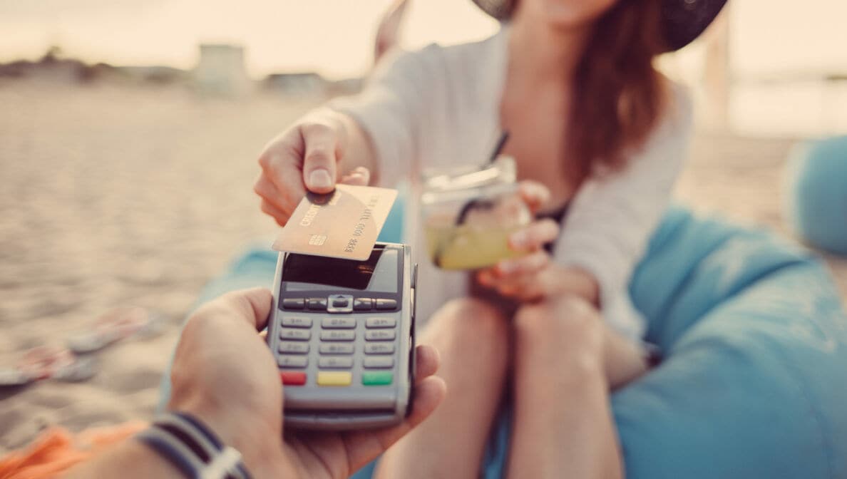 Eine Frau mit Sonnenbrille, Hut und Drink in der Hand am Strand bezahlt per Kreditkarte an einem mobilen Zahlterminal