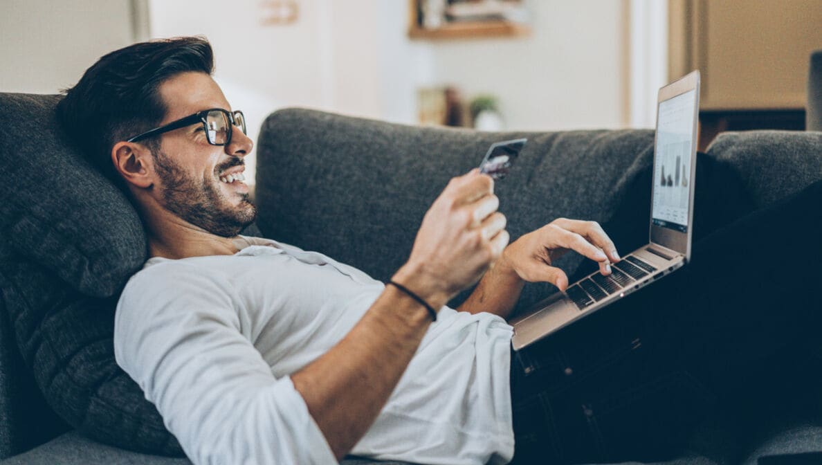 Dunkelhaariger Mann mit Brille liegt mit einem Laptop auf dem Schoß auf einem Sofa und hält in einer Hand eine Bankkarte