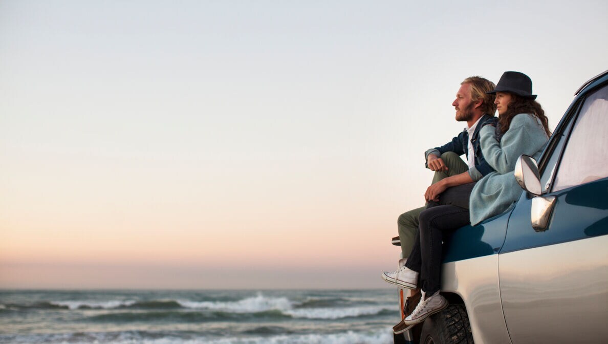 Ein Paar am Meer auf der Kühlerhaube eines Autos mit Brandung im Hintergrund
