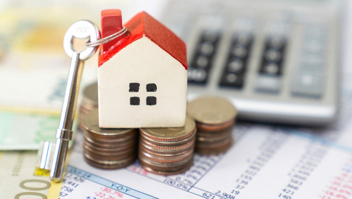 Ein Schlüssel mit Hausanhänger auf einem Stapel Geldstücken stehend. Daneben Geldscheine, ein Taschenrechner und eine Kalkulationstabelle