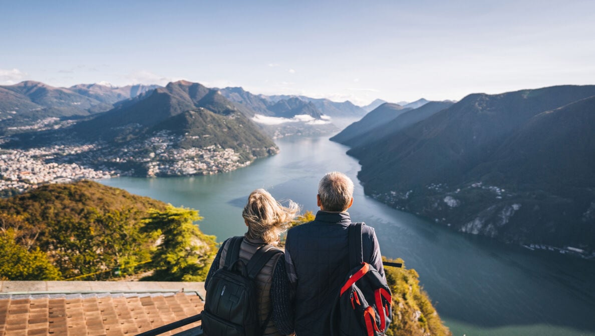 Ein älteres Paar mit Rucksäcken steht auf einer Aussichtsplattform und blickt auf einen Fluss zwischen den Bergen.