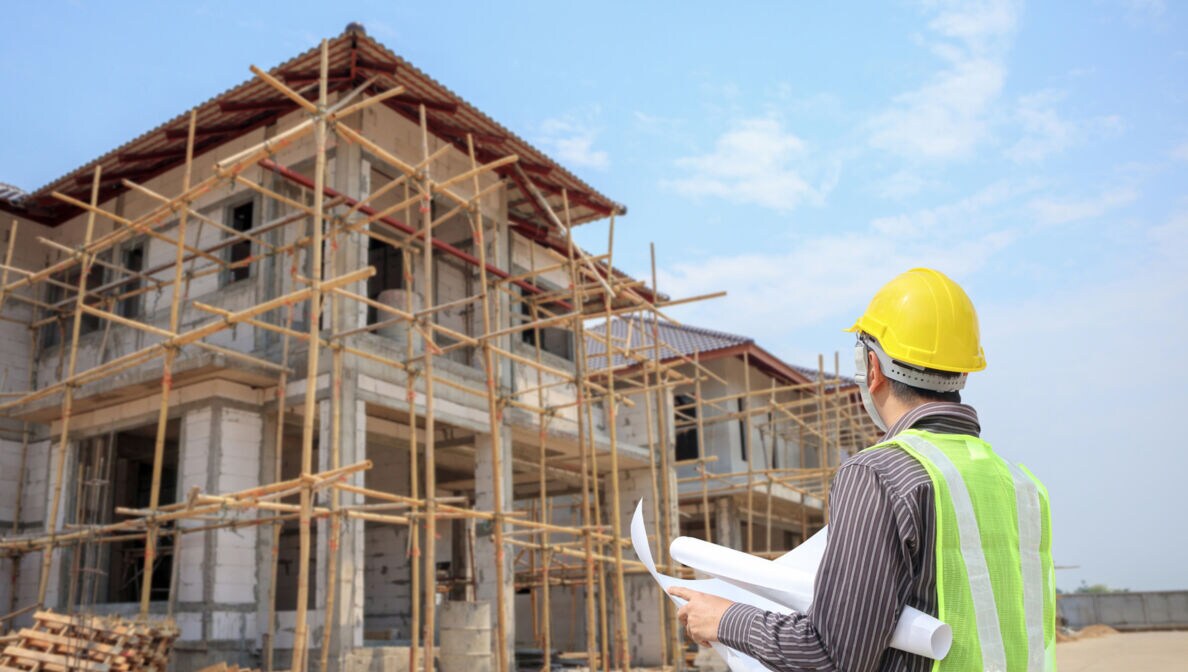 Ingenieur mit gelbem Schutzhelm steht vor einer Hausbau-Baustelle und überprüft seine Pläne