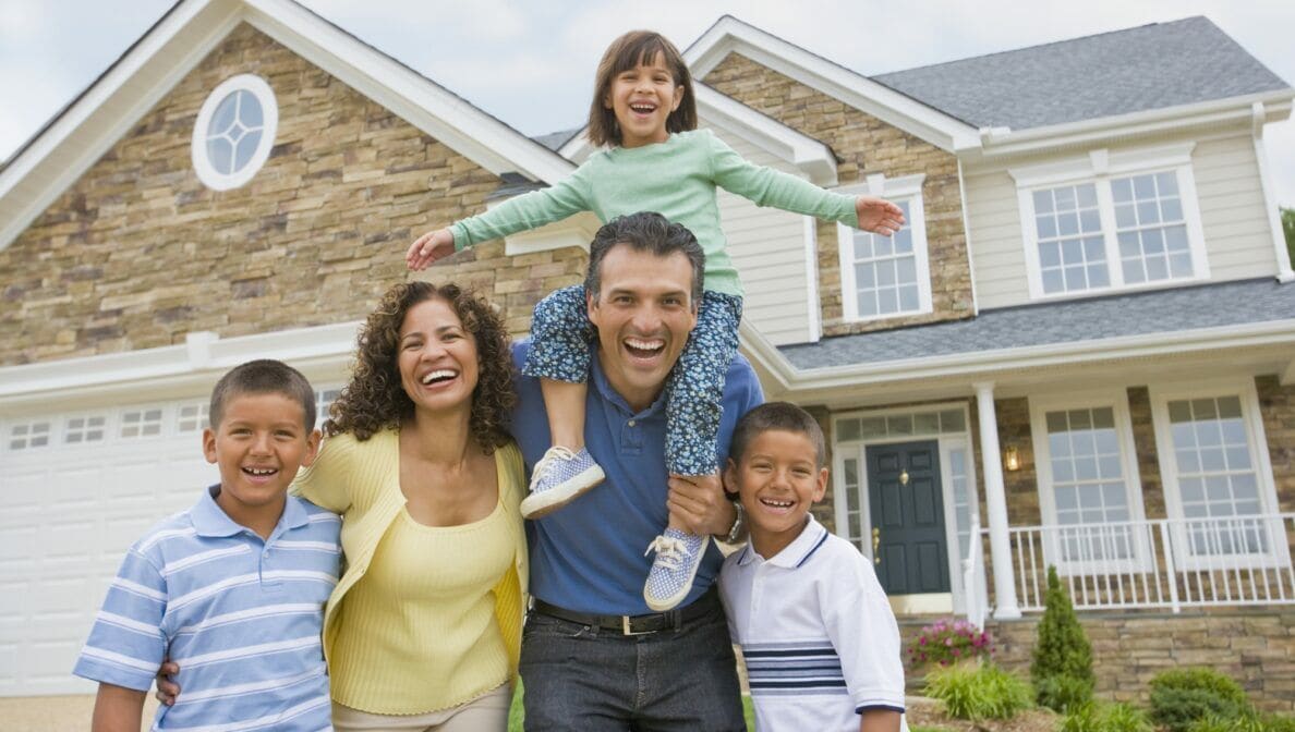 Eine Frau, ein Mann und drei Kinder, eins davon auf den Schultern des Mannes, lachen in die Kamera. Im Hintergrund steht ein Einfamilienhaus.