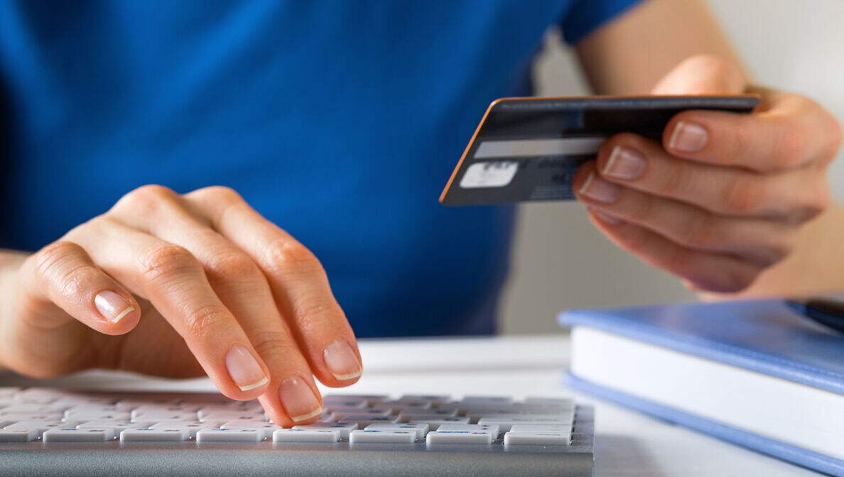Eine Person hält eine Kreditkarte in der Hand und tippt mit der anderen Hand Daten auf einer Tastatur ein