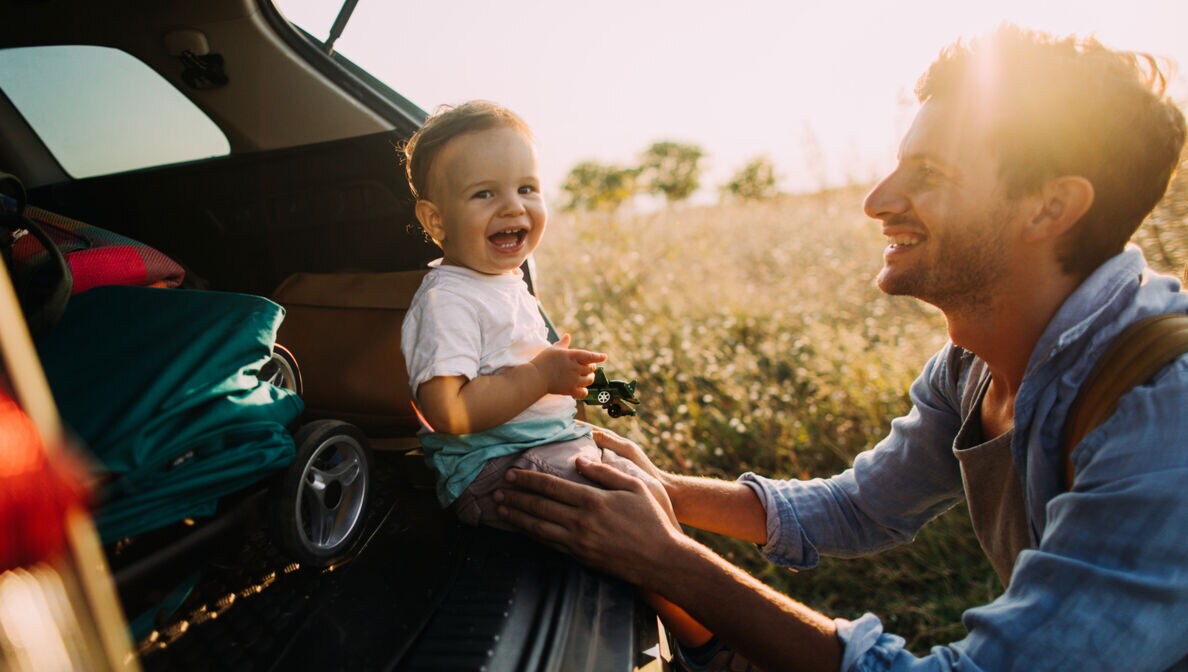 Ein junger Vater hockt vor dem offenen Kofferraum seines Autos, in dem sein lachender Sohn im Babyalter sitzt, während im Hintergrund ein sommerliches Feld zu sehen ist