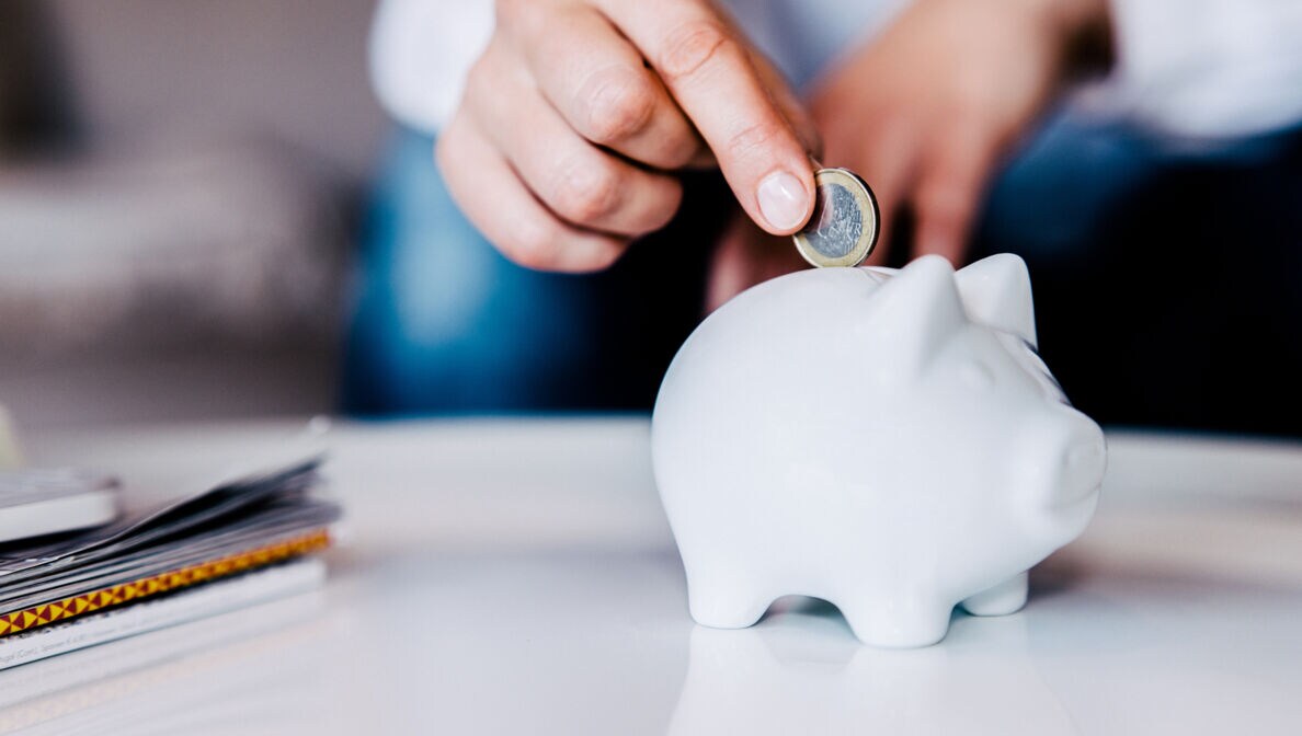 Nahaufnahme der Hand einer Person, die eine Münze in ein weißes Sparschwein wirft