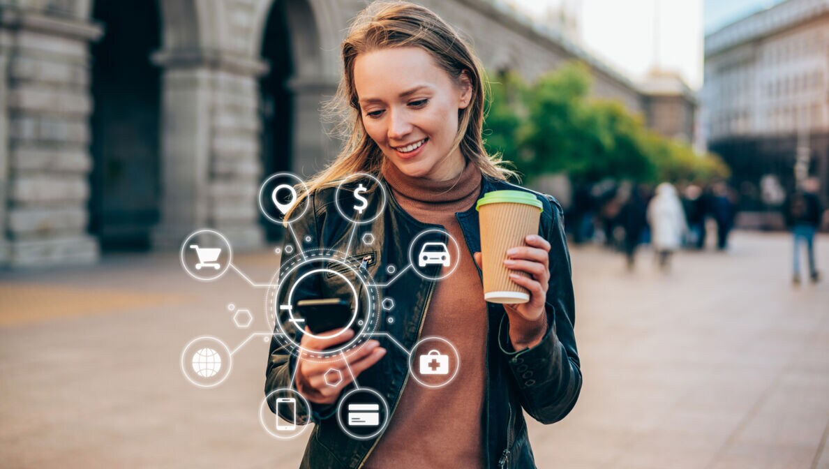 Junge Frau in der Stadt mit Kaffeebecher in der einen und Smartphone in der anderen Hand