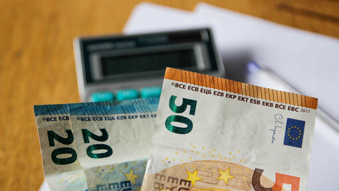 Nahaufnahme mehrerer Geldscheine, im Hintergrund Taschenrechner, Stift und Papier auf einem Schreibtisch