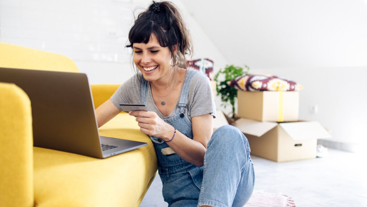 Eine junge Frau mit Kreditkarte in einer Hand vor einem gelben Sofa, auf dem ein Laptop steht