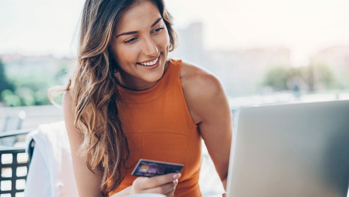 Eine lächelnde Frau auf einer Terrasse vor einem Laptop, in der Hand eine Kreditkarte
