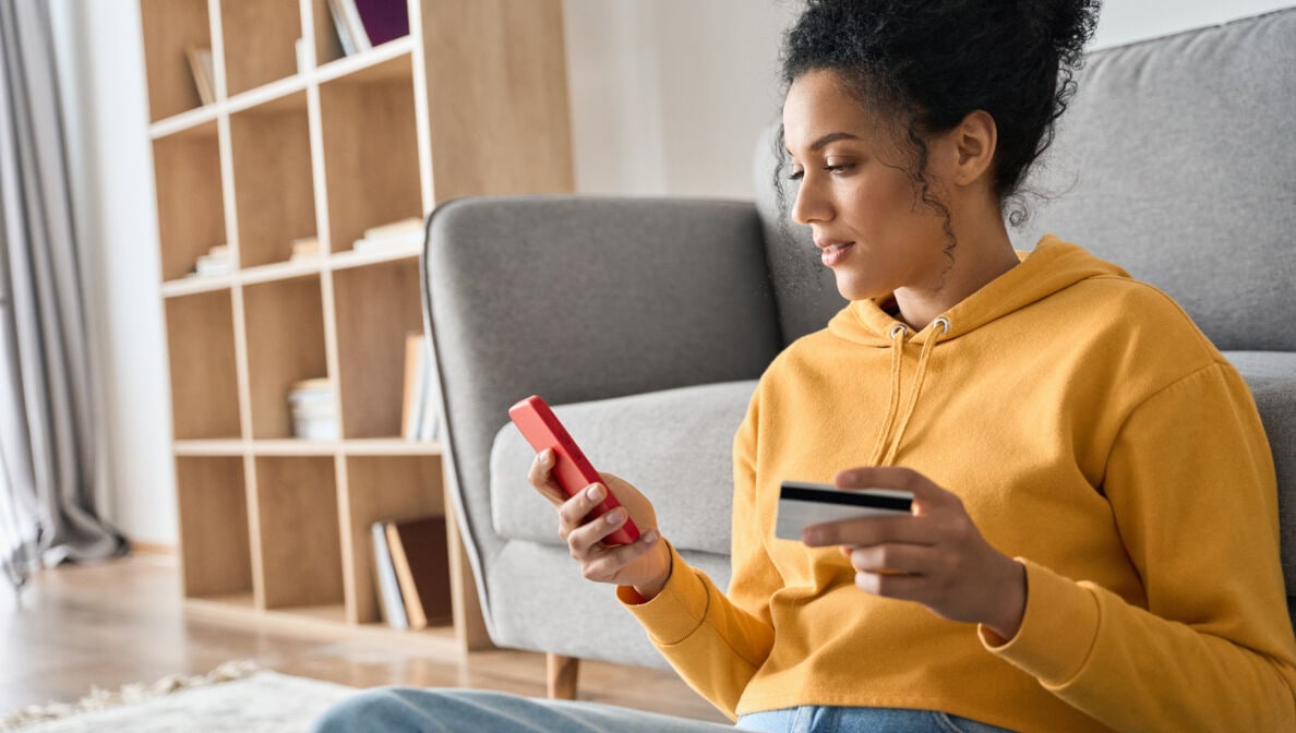 Junge Frau mit gelbem Pullover an ein Sofa gelehnt mit Kreditkarte in der einen und Smartphone in der anderen Hand