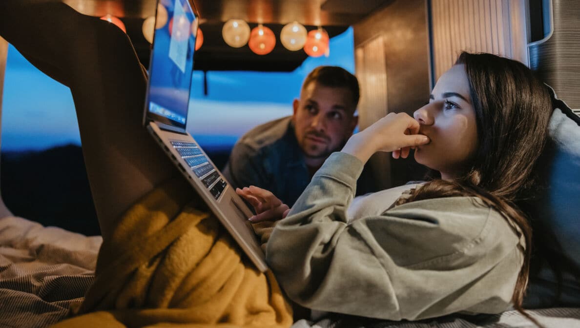 Junge Frau bei Dämmerung in einem offenen Van mit Laptop auf den Beinen und ein Mann, der ebenfalls auf den Bildschirm blickt