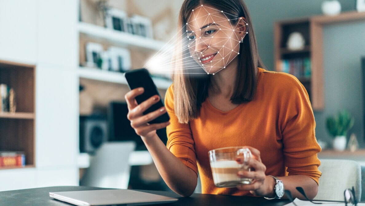 Eine Frau mit Kaffeetasse in der einen Hand und in der anderen ein Smartphone, das Gesichtserkennung durchführt