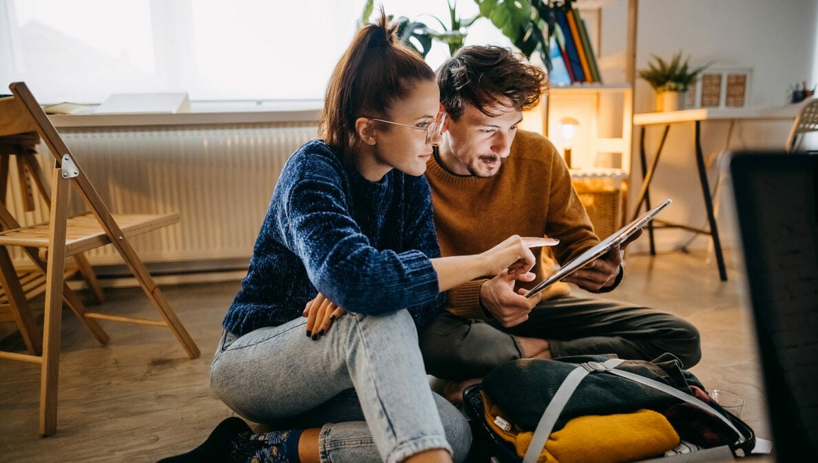 Eine junge Frau und ein junger Mann, in einer Wohnung auf dem Fußboden sitzend, die auf ein Tablet schauen, das er in den Händen hält