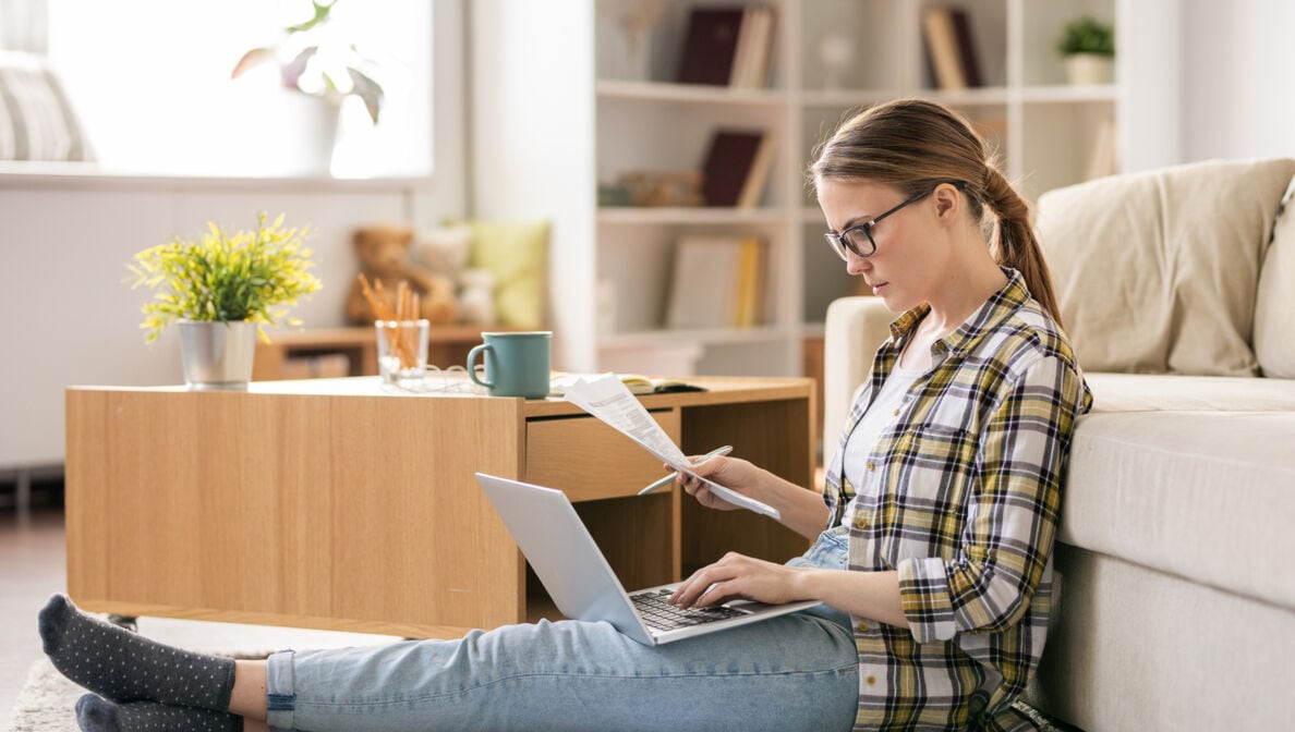 Eine junge Frau sitzt auf dem Boden vor ihrem Sofa, hält einen Zettel in der Hand und schaut auf ihr Laptop.