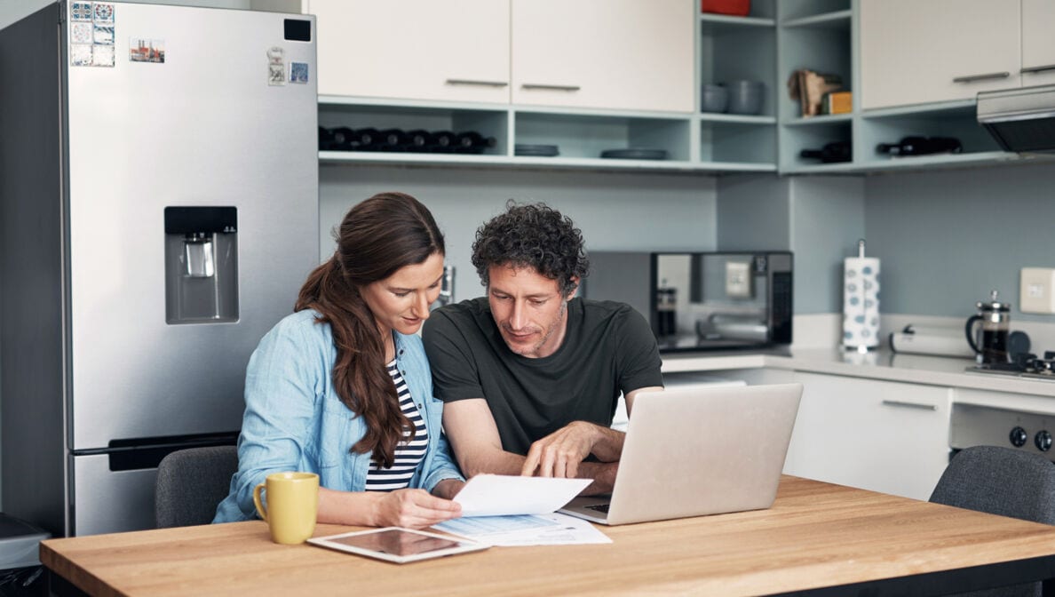 Ein junges Paar sitzt in der Küche mit einem Laptop, einem Tablet und einigem Papierkram.